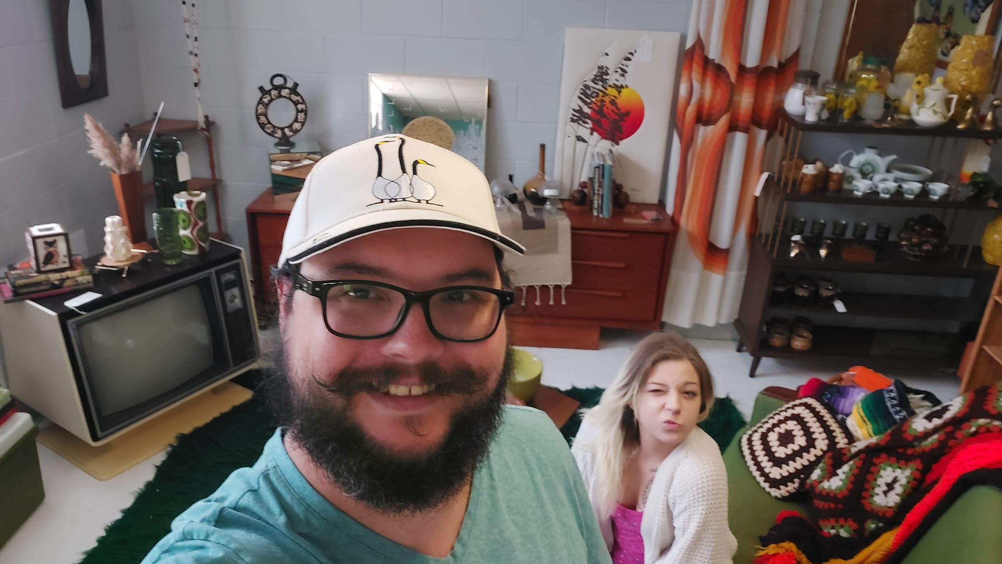 A man with short hair and bushy facial hair stands in an antique store recreation of a 70s living room, a blond woman is sitting on the couch